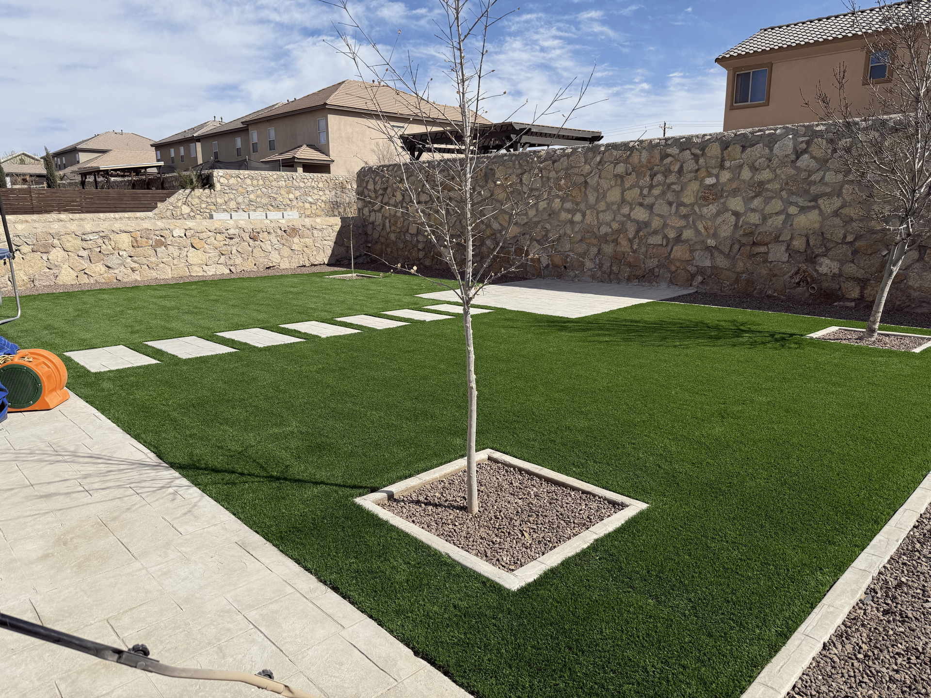 Small backyard with artificial grass, stepping stones, a young tree, and stone walls under a blue sky