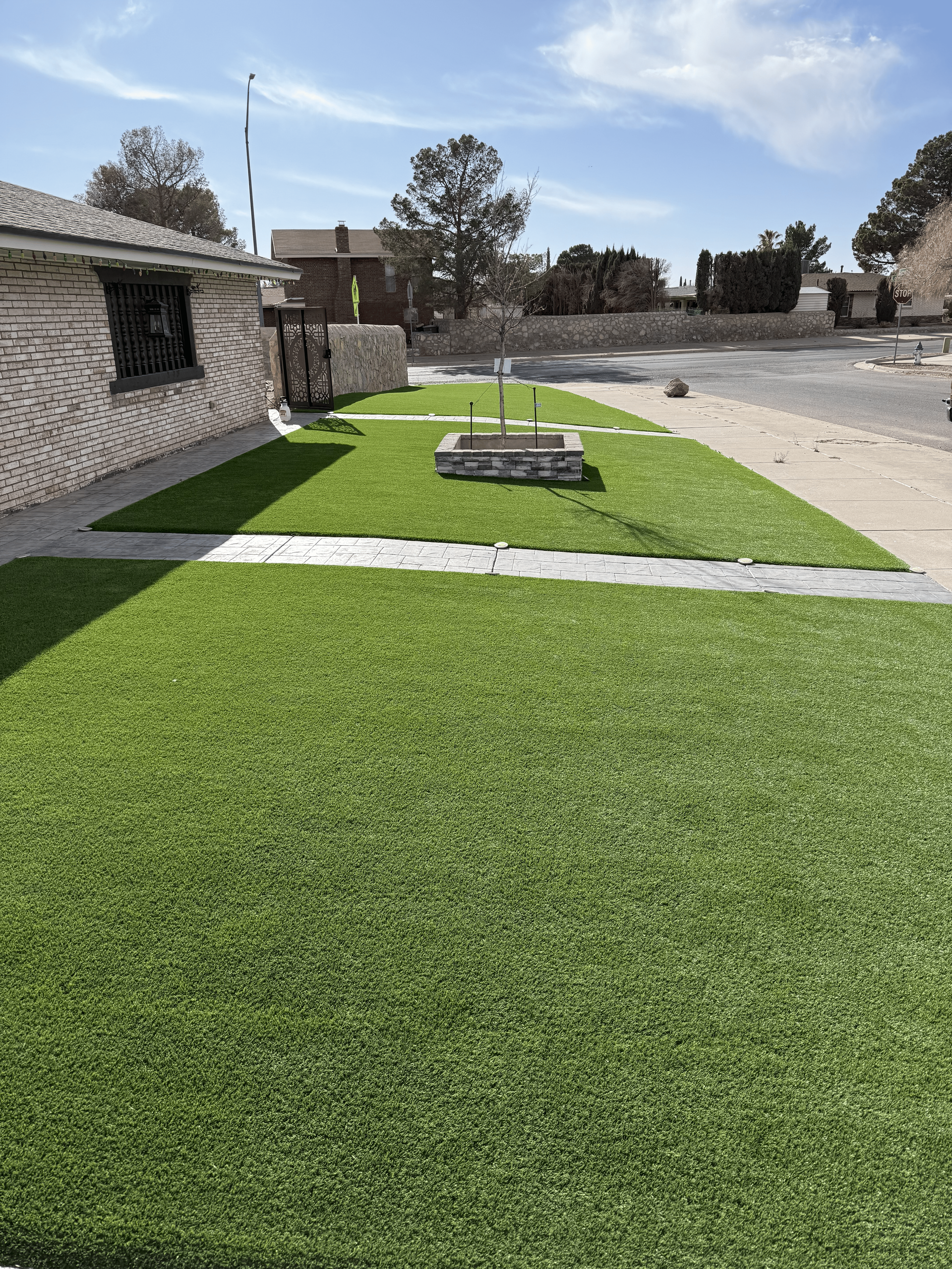 Artificial turf courtyard with a stone planter, walkways, and a brick wall under a blue sky