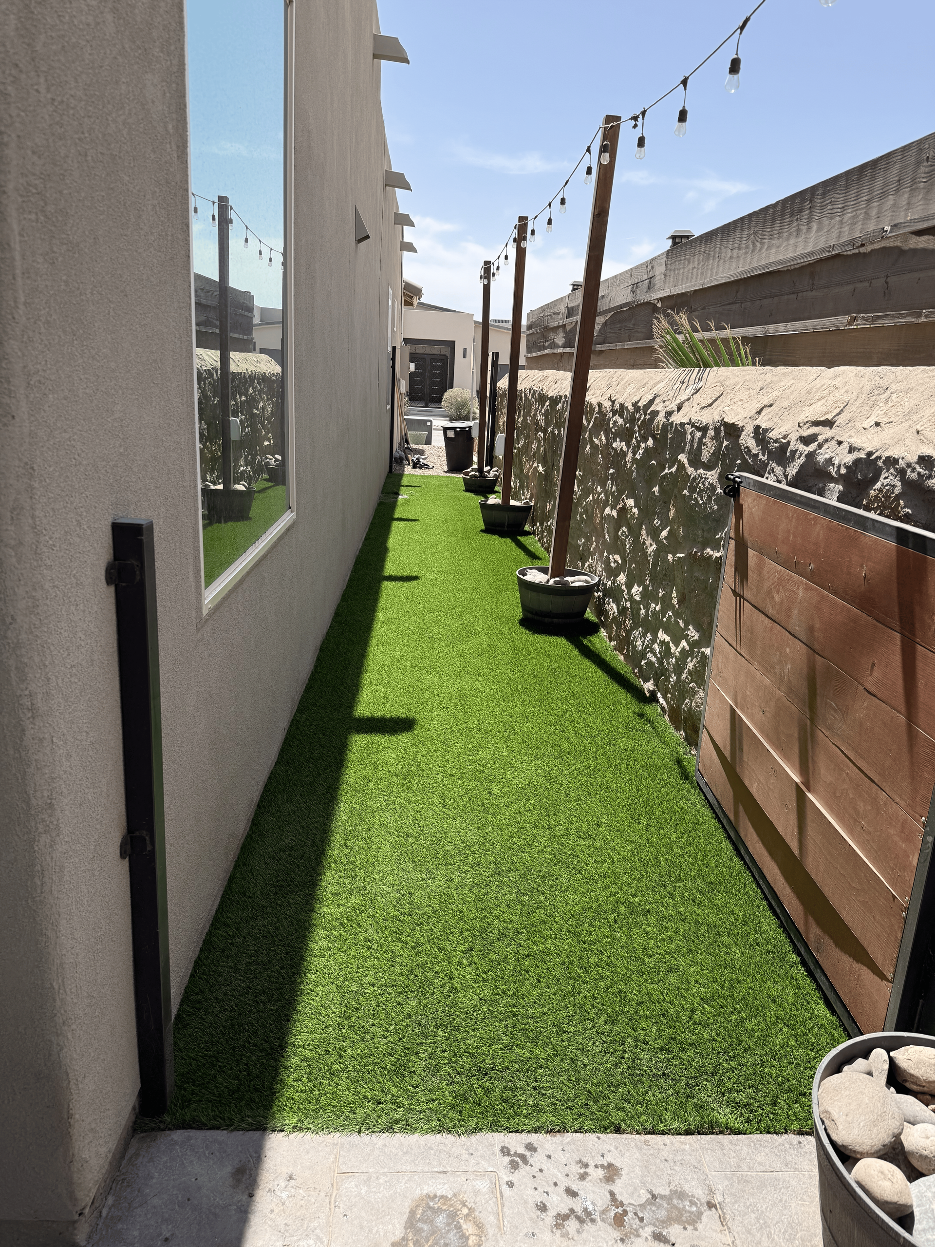 Narrow artificial grass walkway between white and stone walls, with string lights overhead and potted plants.