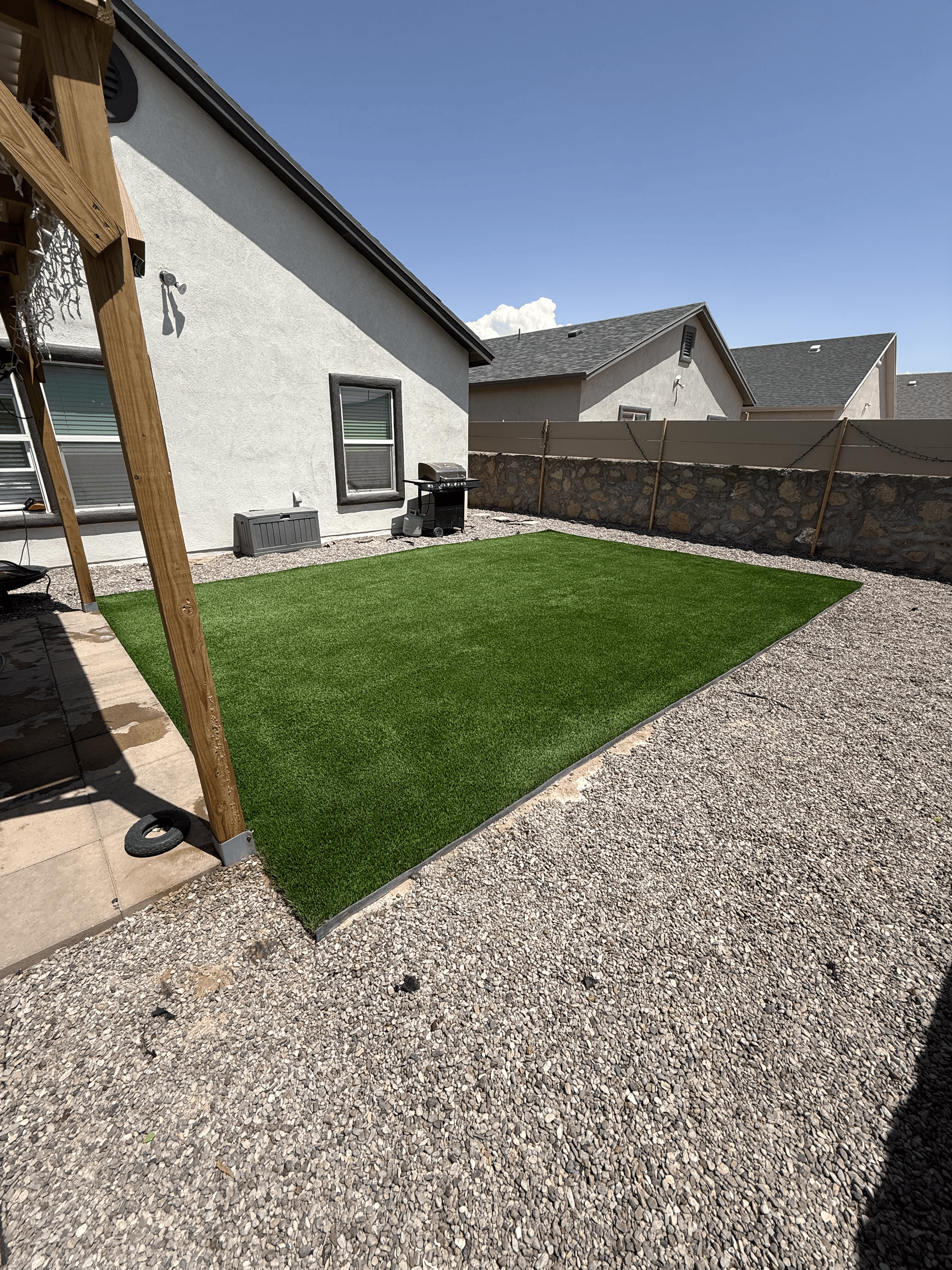Small backyard with a patch of grass, gravel border, white house wall, and stone privacy wall under bright sun
