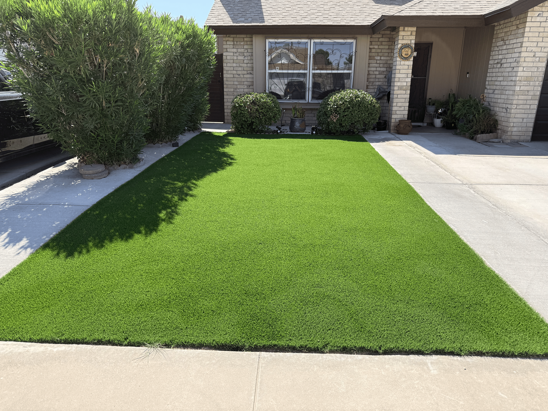 Front yard with bright green lawn, concrete walkways, and a brick house with a shaded porch
