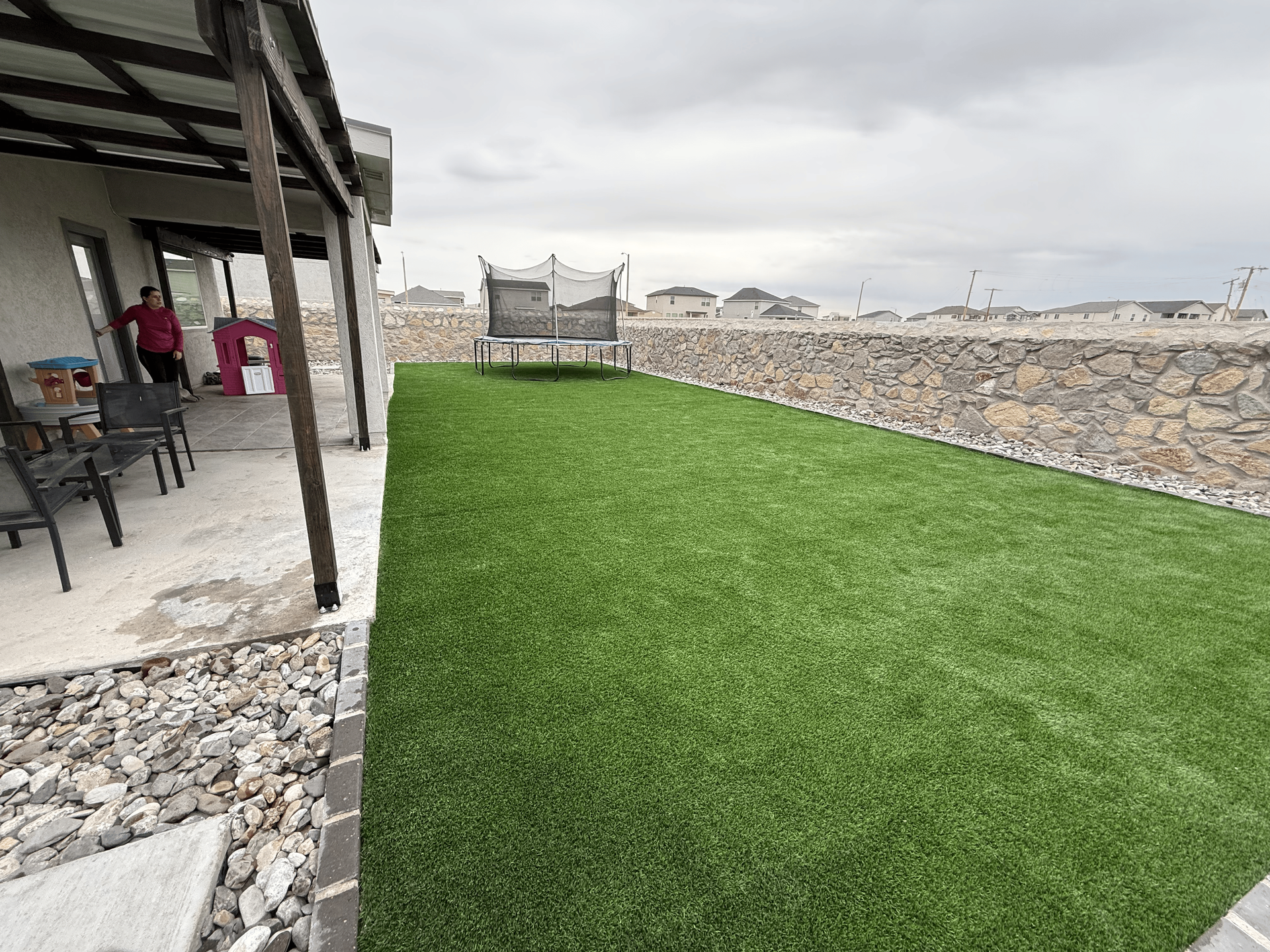 Covered patio beside a green lawn, with a stone wall and outdoor seating under an overcast sky.