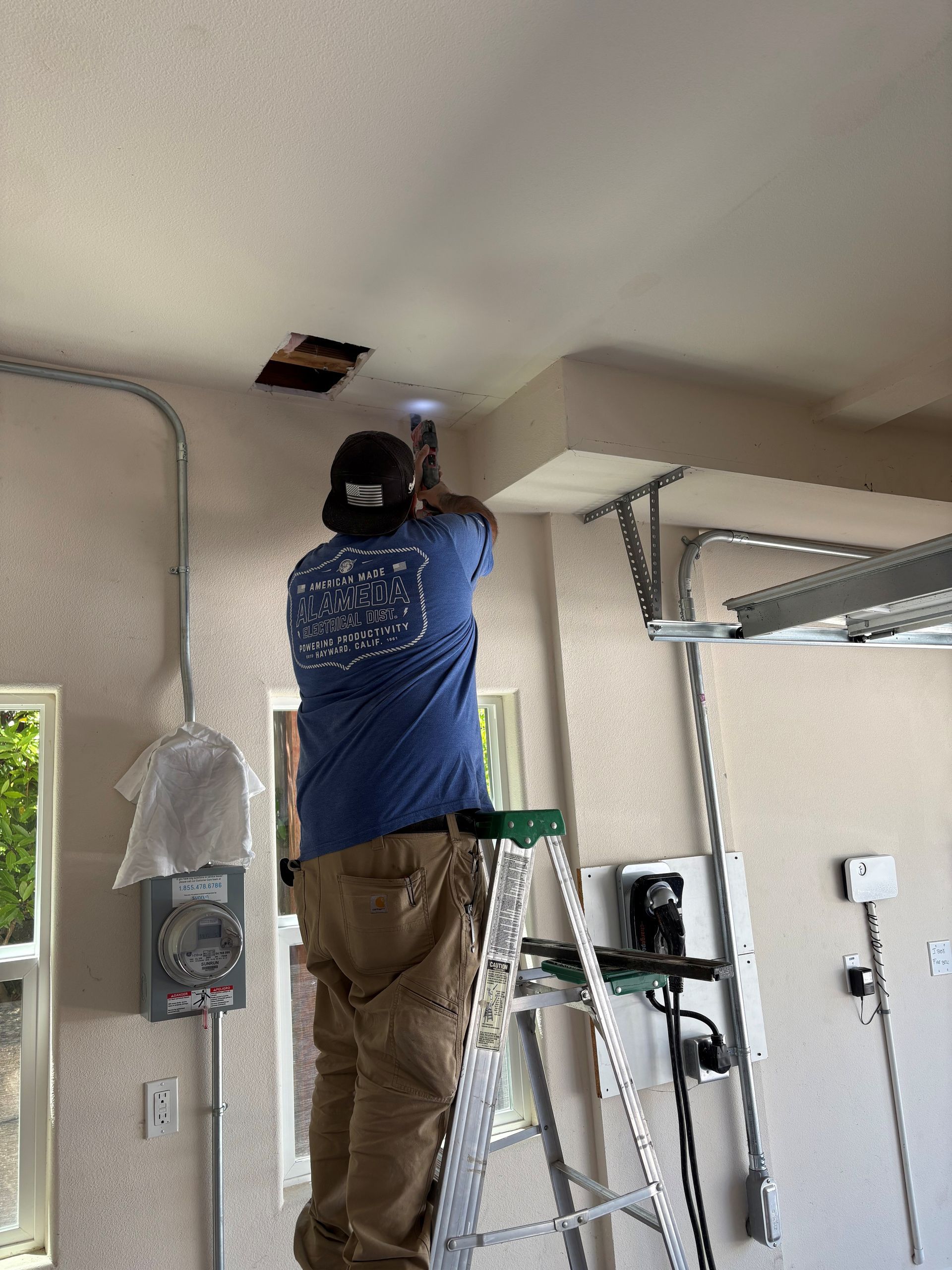 Worker on a ladder installing a ceiling fixture in a garage, with tools and shelves nearby.