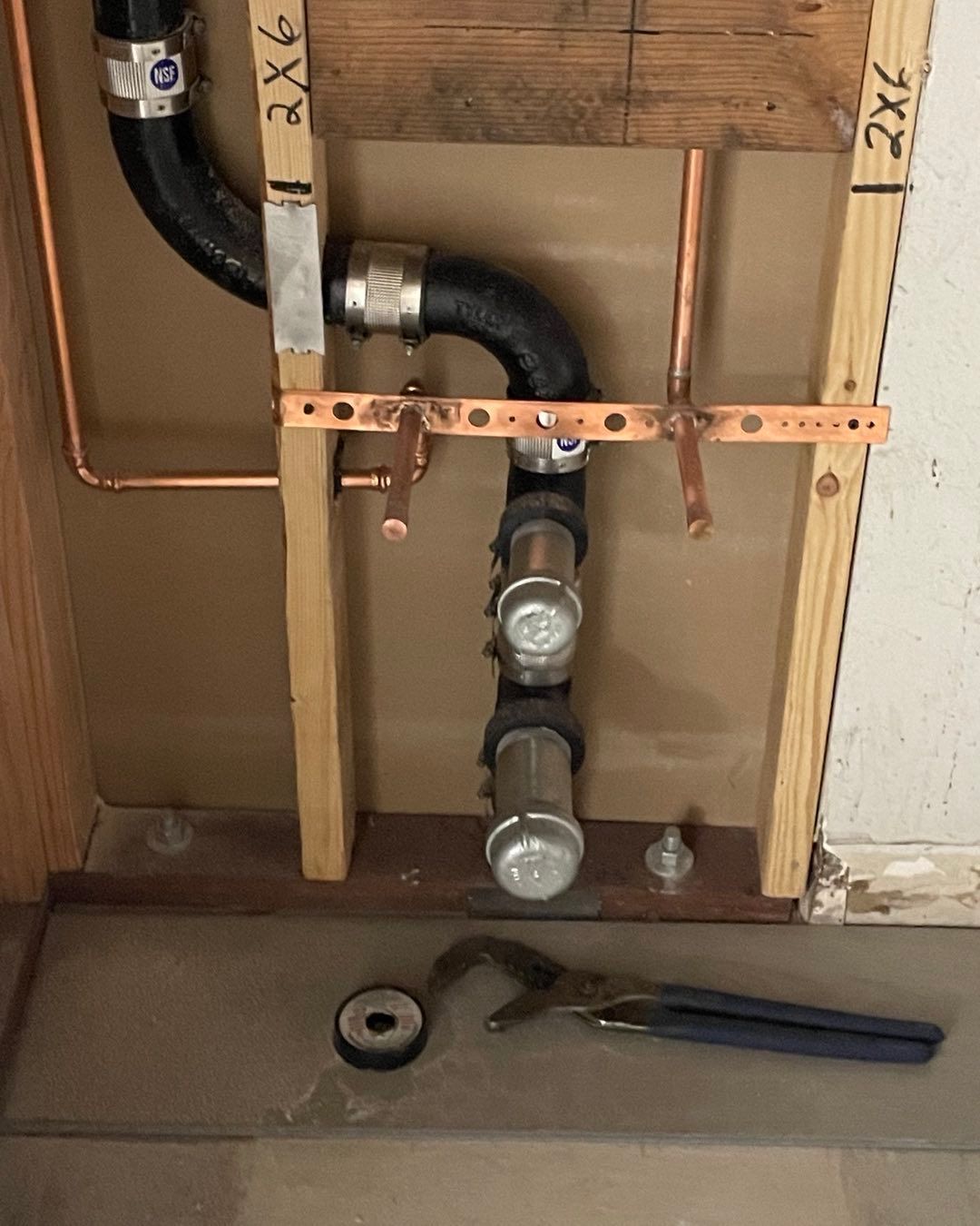 Under-sink plumbing with copper pipes, black drain pipes, wooden framing, and tools on the floor