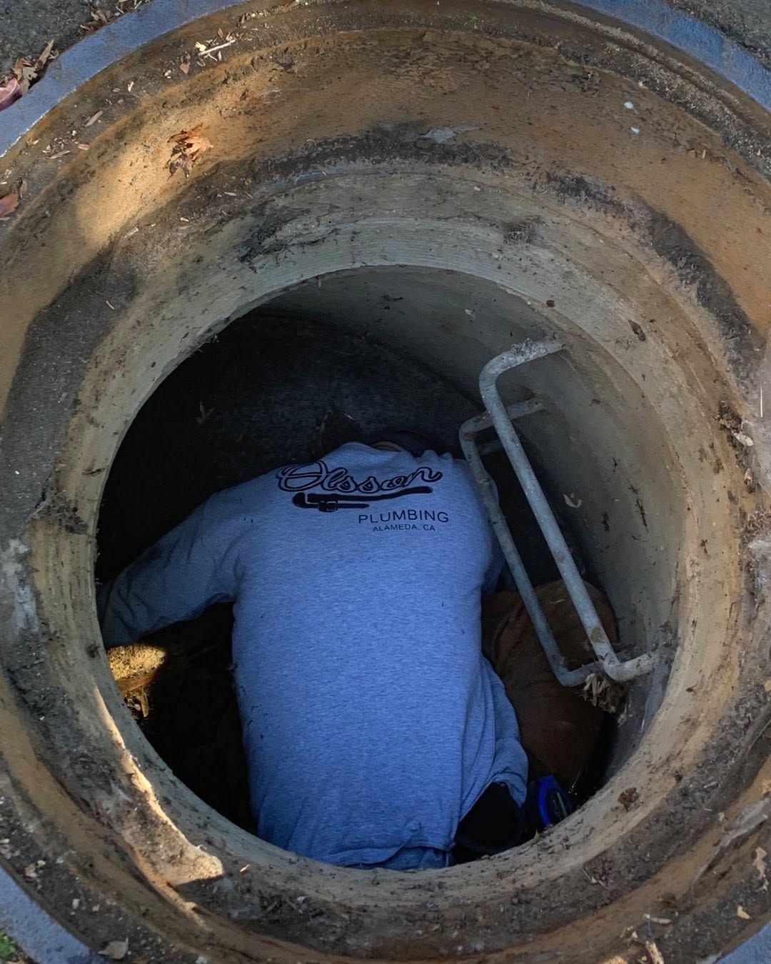 Worker descending into a manhole through a circular opening, wearing a blue shirt and using a ladder