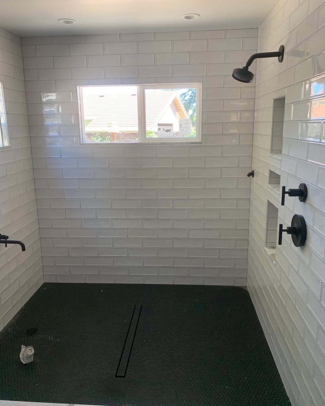 Empty white-tiled shower with black floor mats, window, and wall-mounted showerheads.