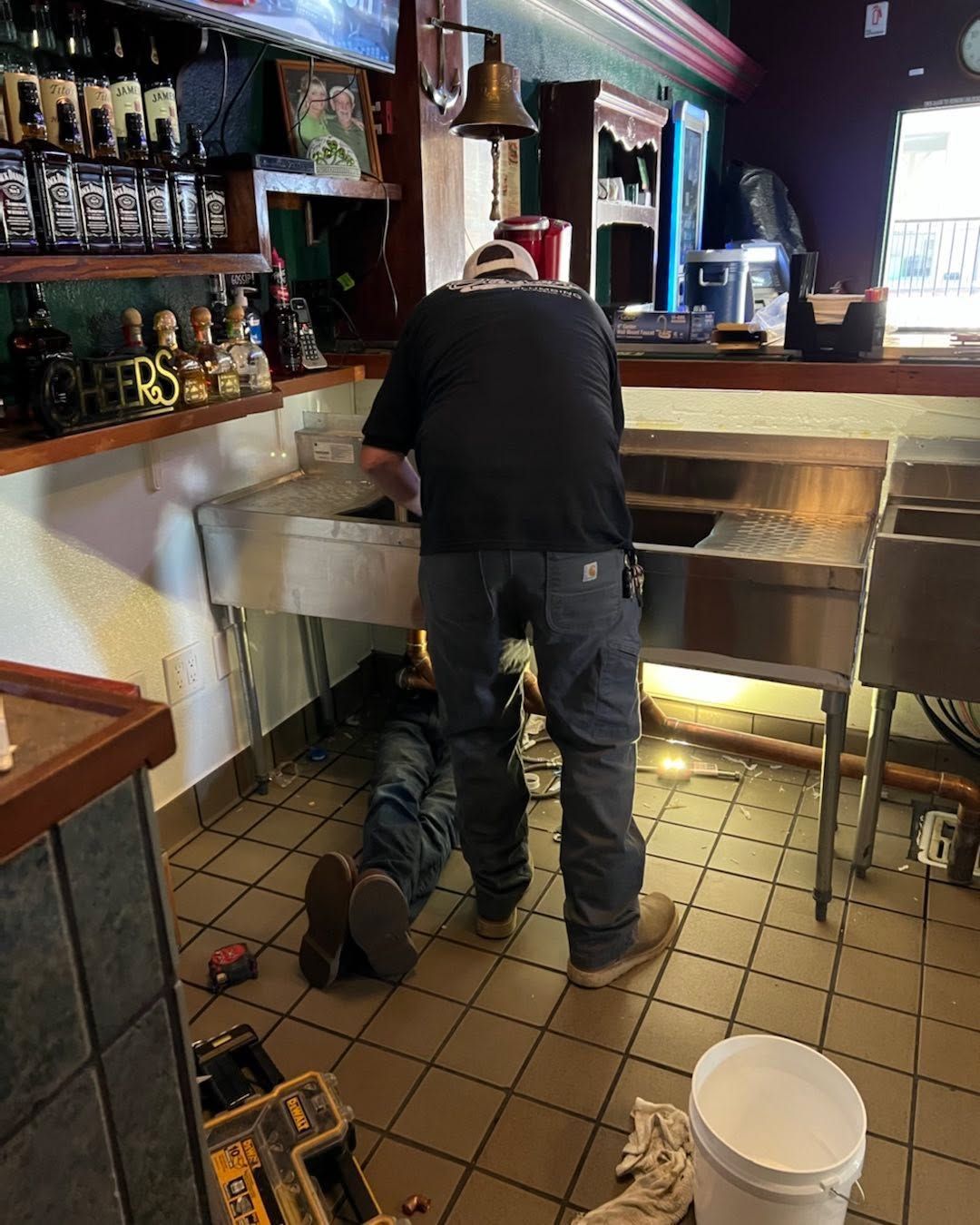 Person working at a bar sink, with a bucket on the tiled floor and shelves of bottles behind them.