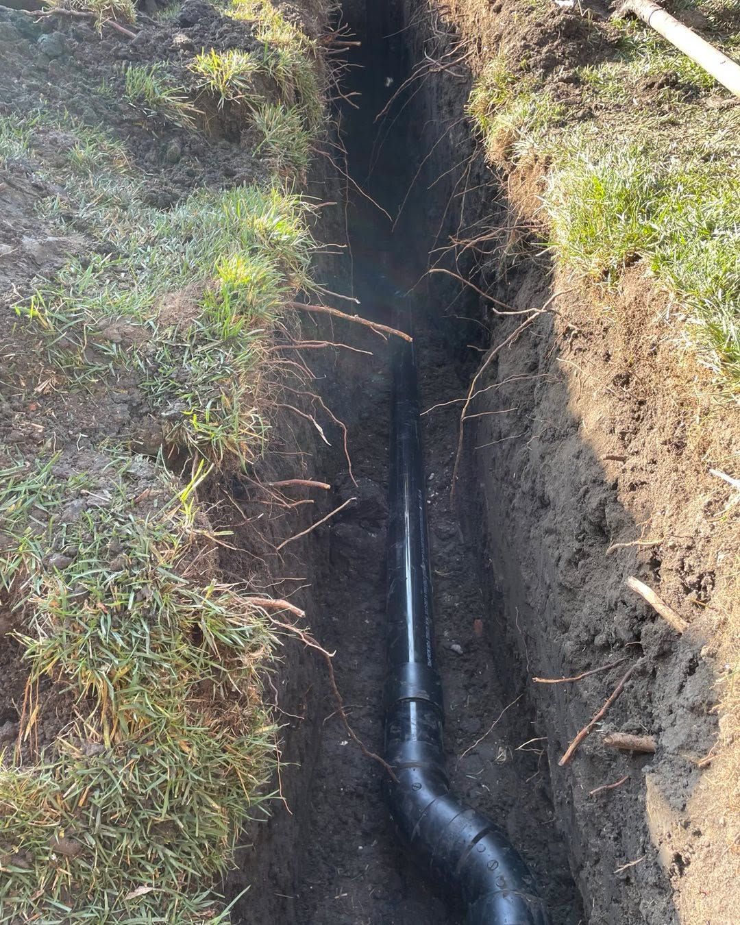 Black pipe in a narrow dirt trench beside grass and soil, with a curved joint near the bottom