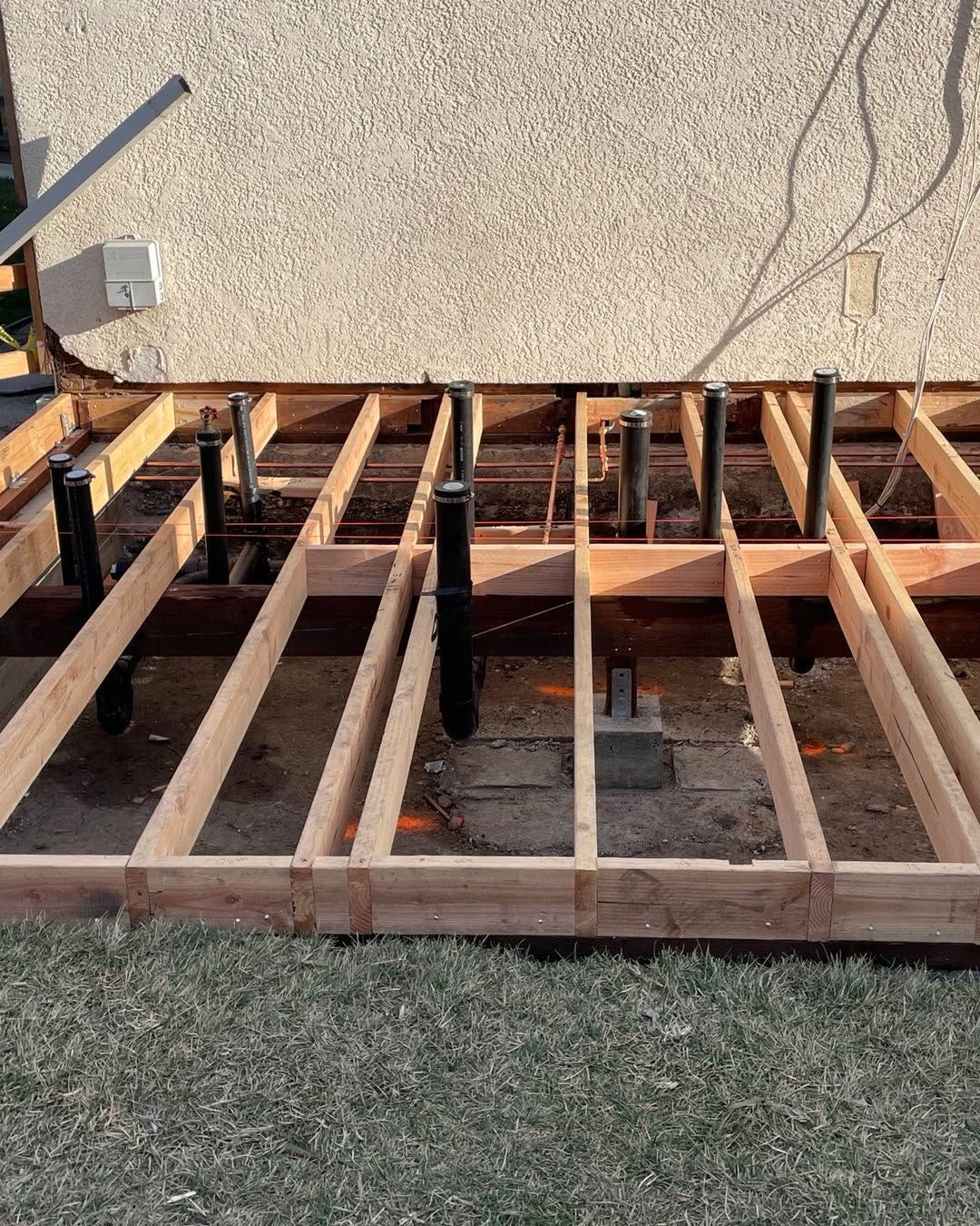 Wooden floor joists and framing laid out over grass beside a stucco wall under construction