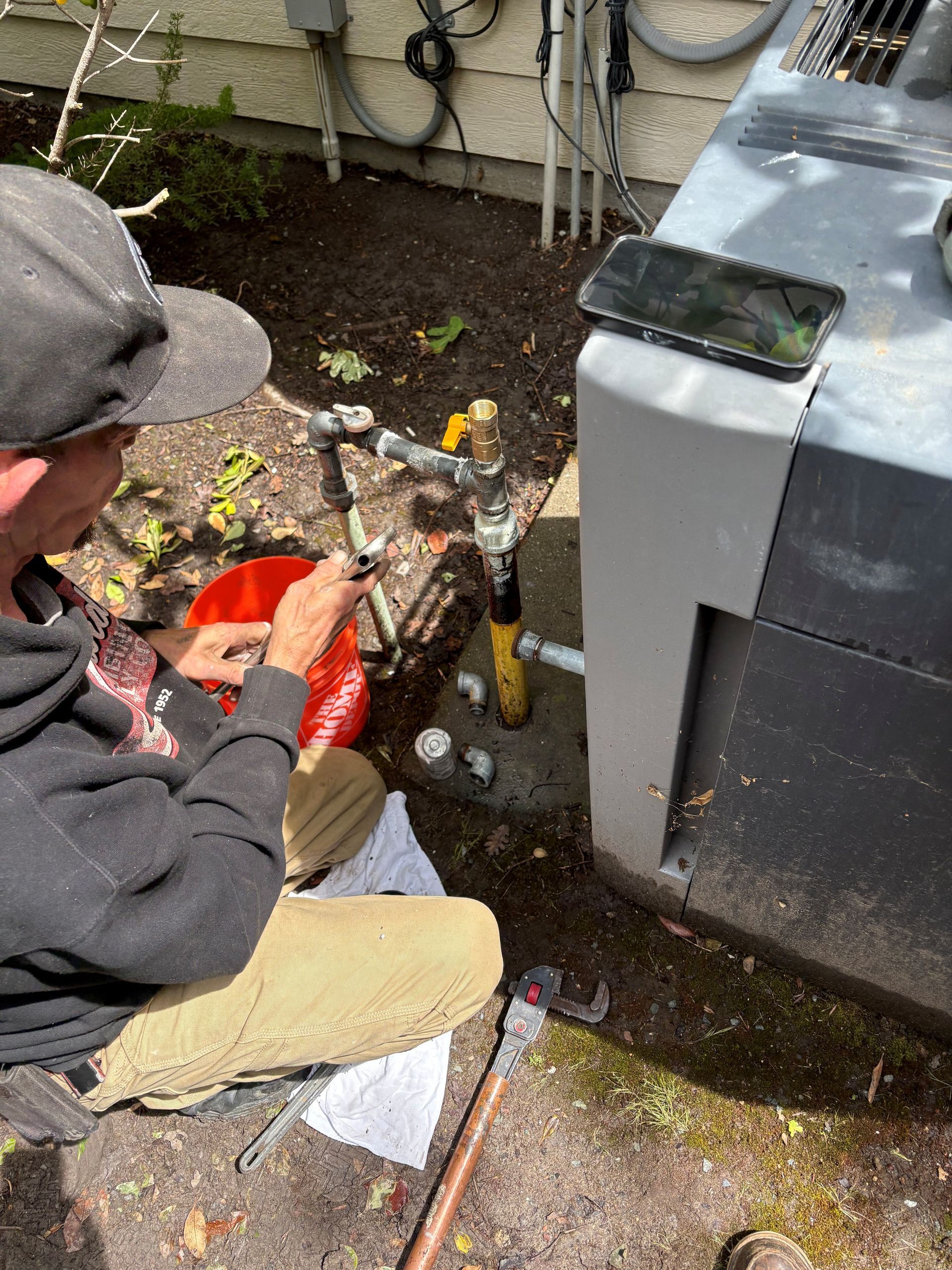 Worker crouches beside outdoor HVAC unit, using a red tool bucket and inspecting yellow pipes.