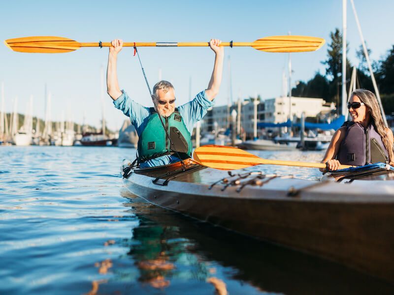 Two people in a canoe on calm water, one raising an orange paddle, with sailboats in the marina behind them