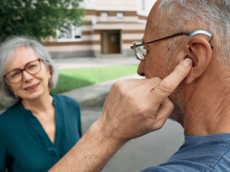 Man pointing to his ear while speaking to an older woman outdoors near a building