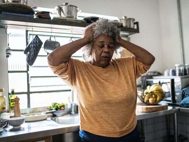 Person standing in a kitchen with hands on head, looking stressed near a countertop with fruit and dishes