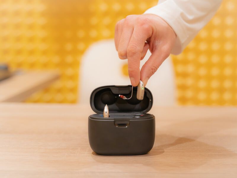 Hand placing a wireless earbud into an open black charging case on a wooden table
