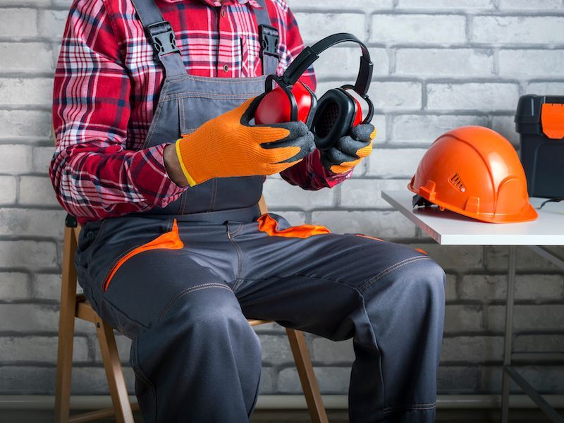 Worker in orange gloves and overalls holding earmuffs, seated beside an orange hard hat in a workshop