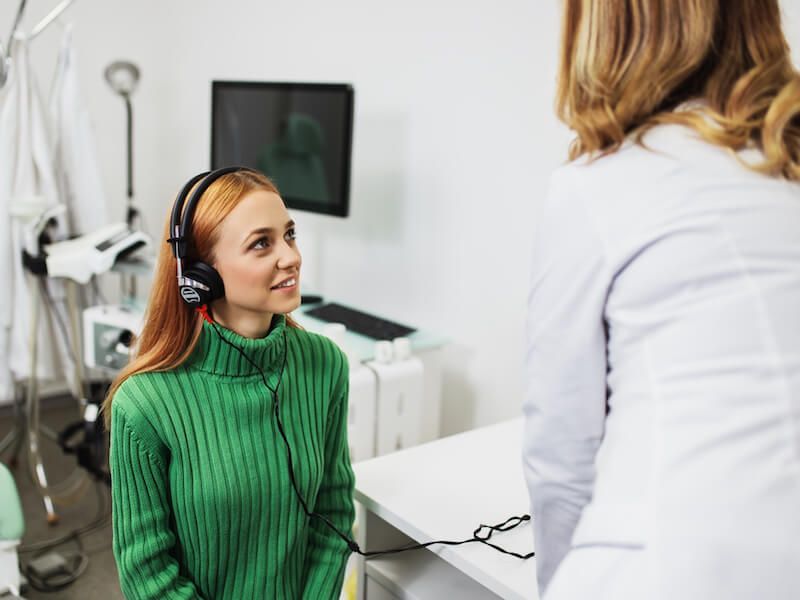 Woman wearing headphones sits in a clinic while a white-coated professional speaks to her