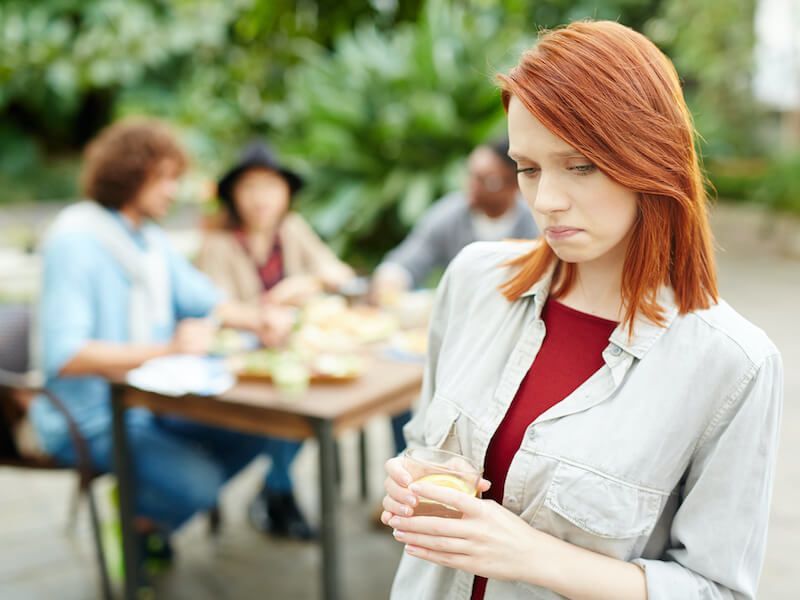 Woman with red hair holding a drink at an outdoor gathering, looking down thoughtfully.