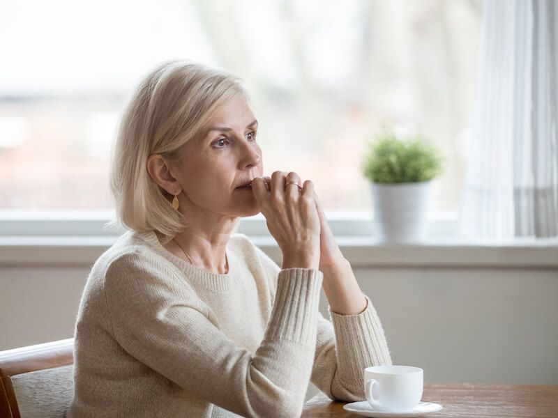 Person sitting by a window, hands clasped near face, looking thoughtfully বাইরে at a small table with a cup.