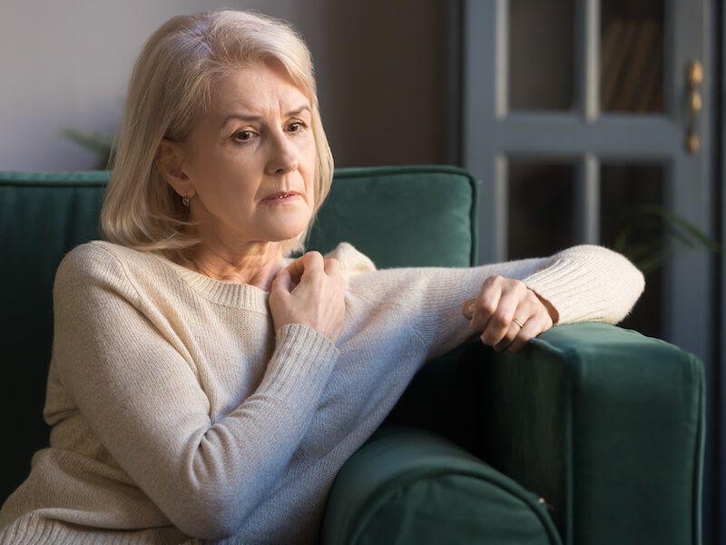 Woman in a cream sweater sitting on a green chair, looking pensive by a window.