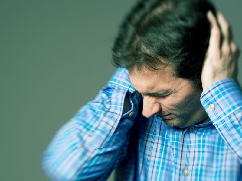 Man in a blue plaid shirt holding his head, looking down with a pained expression