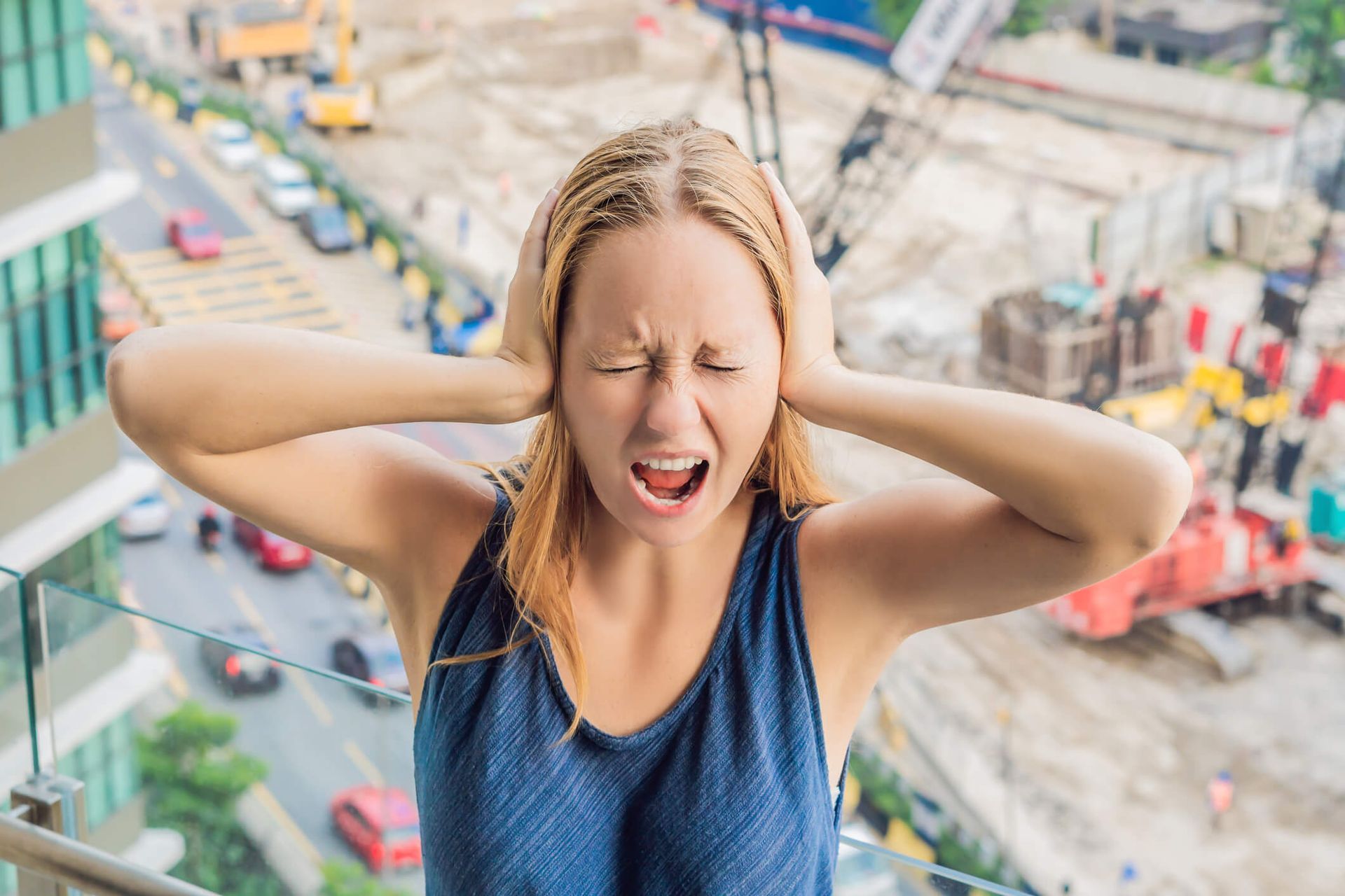 Woman covering ears and grimacing on a glass balcony overlooking a construction site