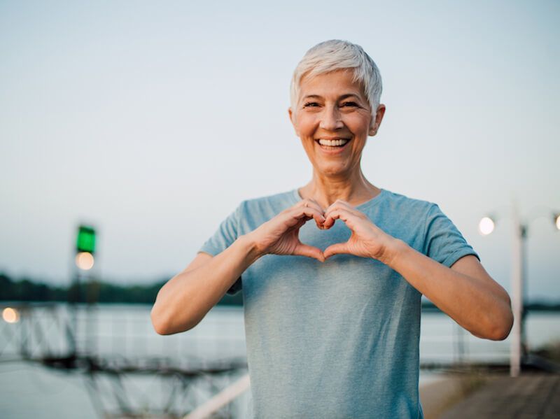 Smiling person in a light blue shirt making a heart shape with hands outdoors at dusk