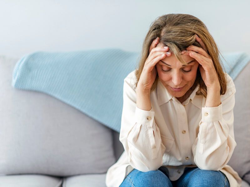 Woman sitting on a couch, holding her head with a worried expression.