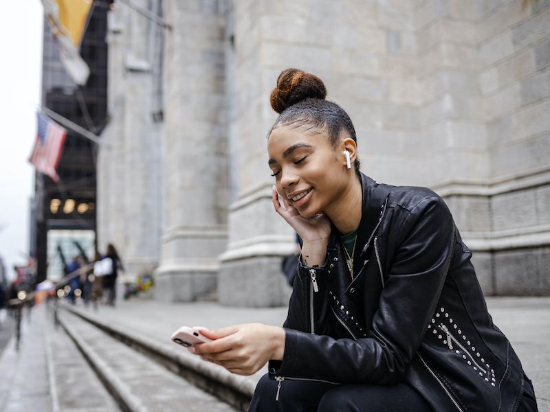 Smiling person leaning on a stone ledge, looking at a phone on a city street.