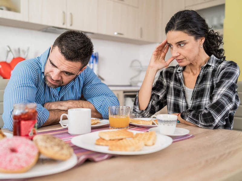 Two people sit at a kitchen table eating breakfast with coffee, pastries, and juice.
