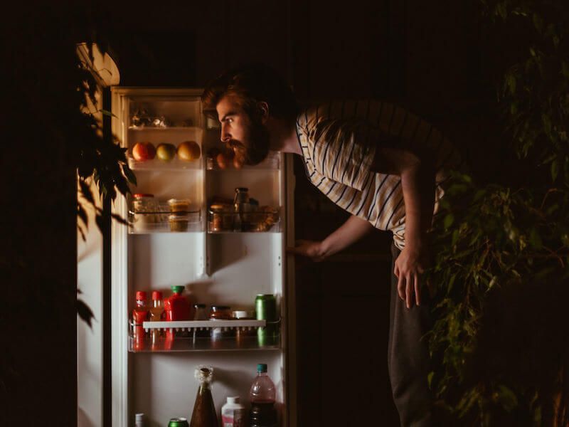 Man peeks into an open fridge with drinks and fruit inside, lit warmly in a dark room