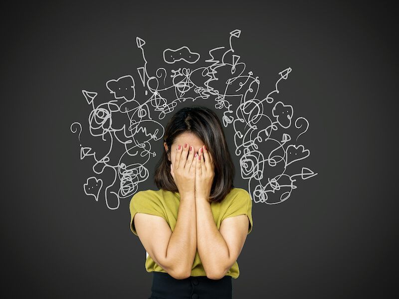 Woman covering her face with hands, standing before a dark background with white doodle scribbles of thoughts above her head