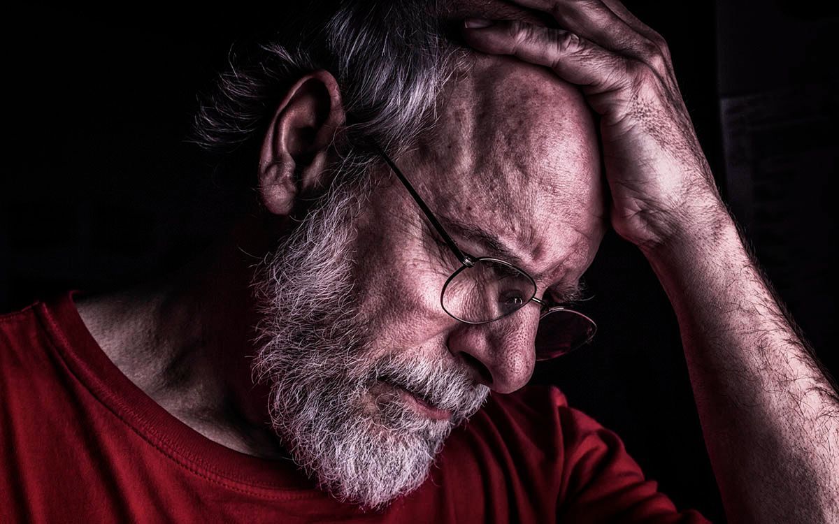 Man with glasses and beard, hand on head, looking down in a red shirt against a dark background