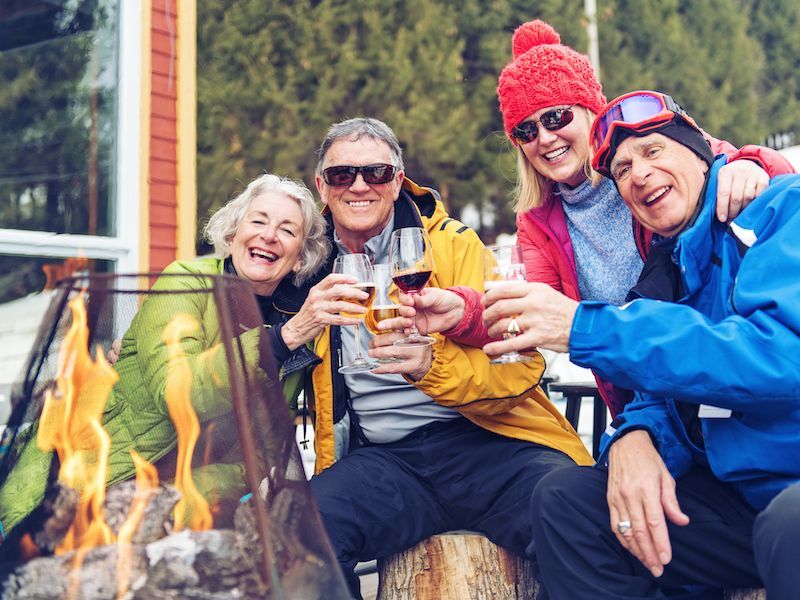 Four friends in winter gear smiling by a campfire, holding drinks outdoors.