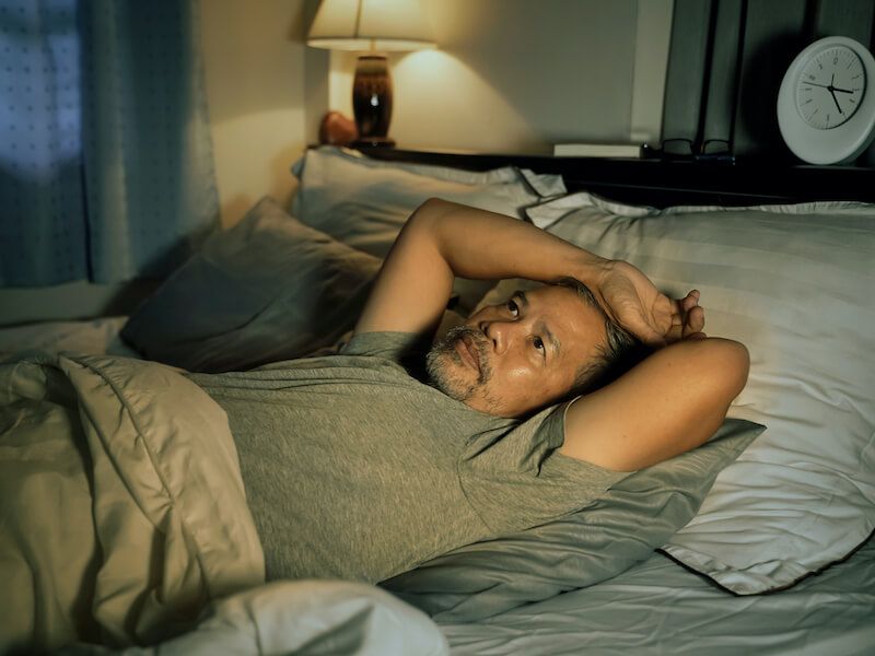 Person lying on a bed with hands behind head, looking tired in a dimly lit bedroom.