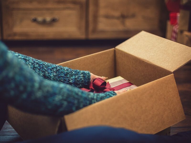 Person packing items into a cardboard box on a table, with red and pink clothing visible in a home setting