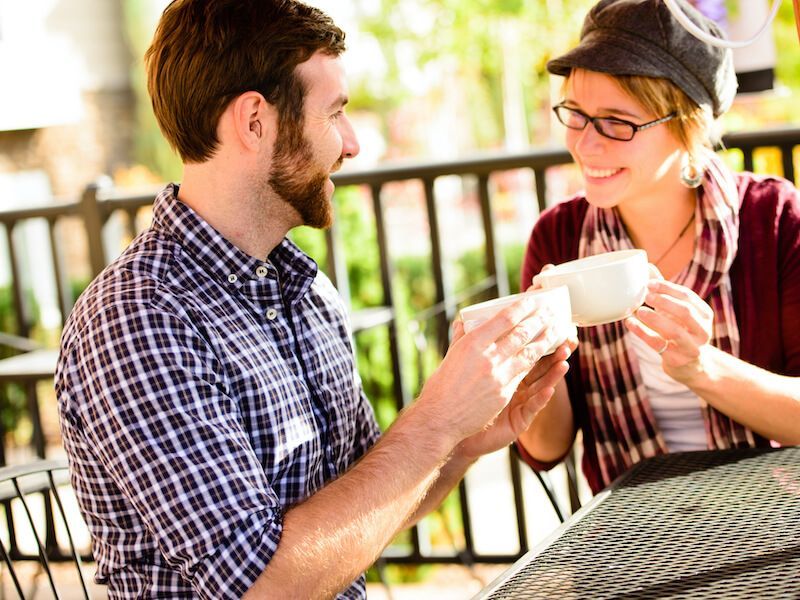 Two people smiling and clinking coffee mugs at an outdoor café table