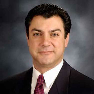 Formal portrait of a man in a dark suit, white shirt, and red tie against a gray studio background