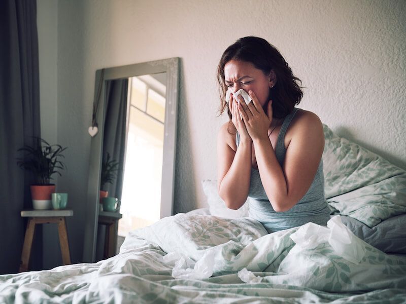 Woman sitting in bed and blowing her nose with tissues in a softly lit bedroom