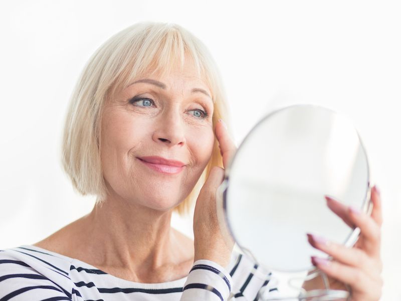 Woman in a striped shirt looking into a handheld mirror and touching her face against a white background