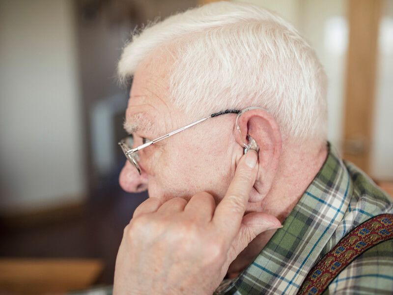 Elderly man with white hair adjusting a hearing aid, seen in profile indoors