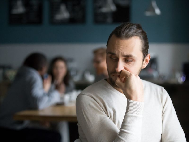 Man in a light sweater resting his chin on his hand in a dim café, looking thoughtful.