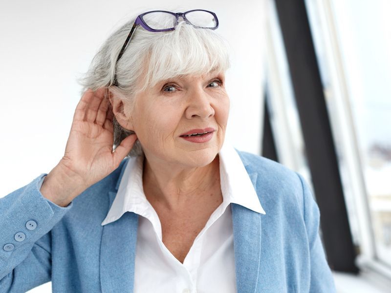 Senior woman in a light blue sweater listening with hand to ear, glasses on her head, indoors by a bright window