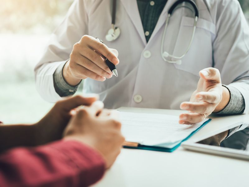 Doctor speaking with a patient across a desk, holding a pen and clipboard in a clinic office