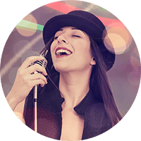 Woman singing into a microphone under colorful lights, wearing a black hat and smiling.