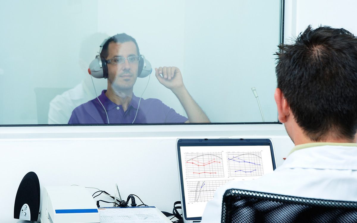 Person wearing a headset speaks through a glass partition to someone at a computer desk.
