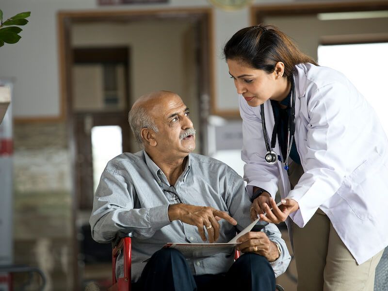Doctor speaking with elderly patient in wheelchair, pointing at his hand in a bright clinic room