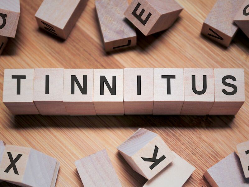 Scrabble tiles spelling “TINNITUS” on a wooden surface, with scattered letter tiles nearby