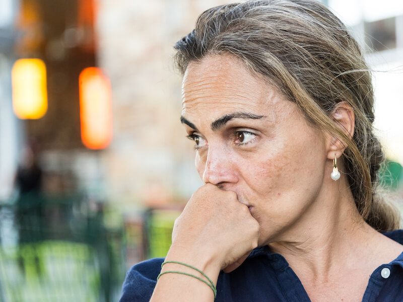 Woman with braided hair resting her hand on her mouth, looking worried outdoors in a blurred urban setting