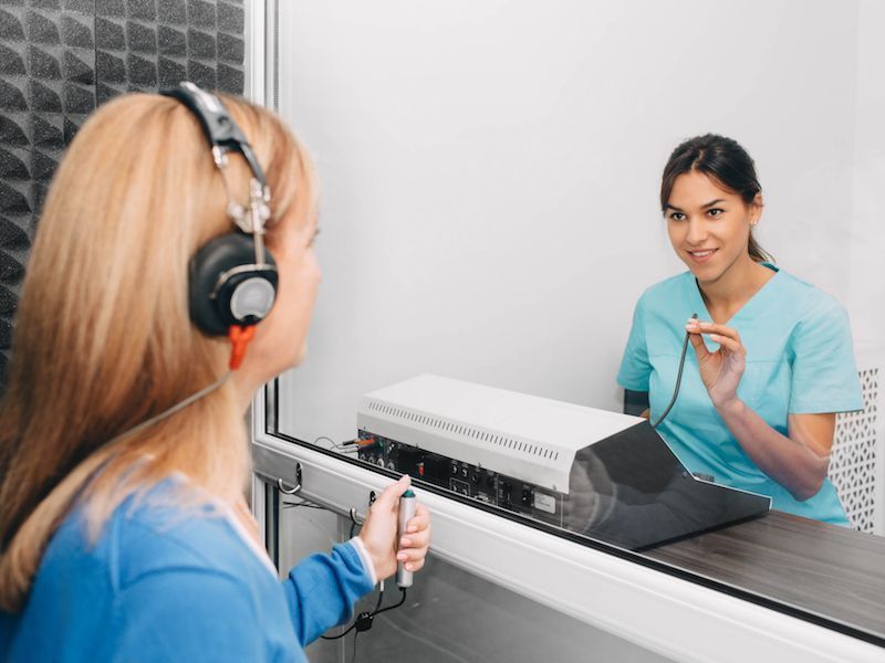 Woman at a clinic counter speaking through a glass barrier to a receptionist in scrubs
