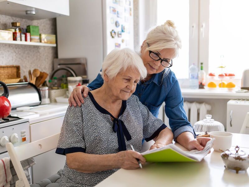 Two women reading a green book together in a bright kitchen